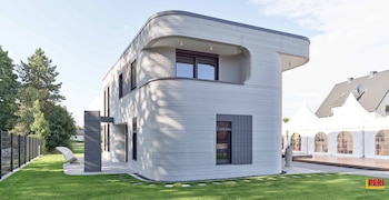 Casa de dos plantas, paredes grises con textura horizontal, ventanas negras, césped verde, camino de losas, valla metálica, árboles, tiendas blancas, cielo azul