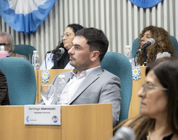 Hombre de traje gris sentado, etiqueta con Santiago Aberastain, Diputado Provincial. Botellas de agua, vaso y micrófono. Bandera argentina y personas al fondo