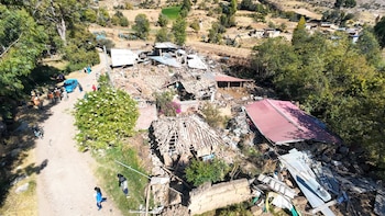 Vista aérea de un camino de tierra con personas y un coche, junto a casas destruidas y edificios dañados en una zona rural con árboles y montañas