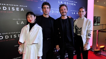 Cuatro personas posan frente a un banner negro con las palabras 'Christopher Nolan' y 'La Odisea'. Dos hombres con trajes oscuros. Una mujer con abrigo blanco, otra con blazer blanco