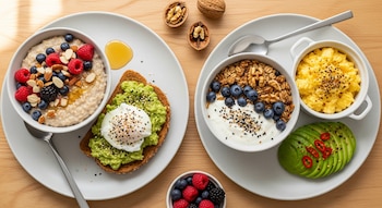 Dos platos de desayuno balanceado con avena, frutos rojos, aguacate, huevo escalfado, yogur, granola, arándanos, huevos revueltos y nueces sobre mesa de madera.