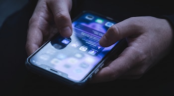 Manos sosteniendo un teléfono inteligente encendido con iconos de aplicaciones en un entorno oscuro.