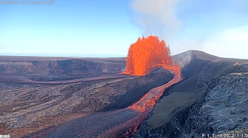 Un volcán expulsa lava incandescente de un cráter con flujos de roca fundida que recorren un paisaje volcánico oscuro, bajo un cielo azul y con un logo USGS