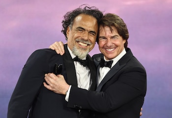 Alejandro González Iñárritu y Tom Cruise sonríen y se abrazan vestidos con trajes de etiqueta negros frente a un fondo de tonos violetas