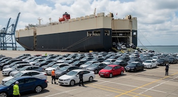 Buque transportador de automóviles anclado en un puerto, descargando vehículos nuevos de diversos colores en el muelle donde hay grúas y personal.