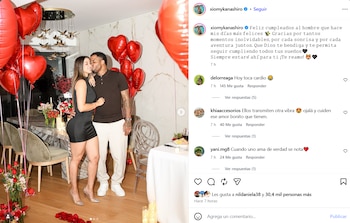 Una pareja se abraza en un comedor decorado con globos rojos en forma de corazón, flores, velas y una mesa servida