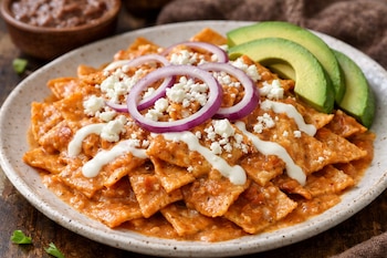 Plato de chilaquiles con totopos cubiertos de salsa naranja, queso blanco desmenuzado, aros de cebolla morada y rebanadas de aguacate.