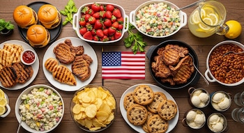 Mesa de madera con hamburguesas, chuletas de cerdo, pechugas de pollo asadas, papas fritas, frijoles, ensalada de papa, fresas, limonada, galletas y helado, bandera de Estados Unidos.