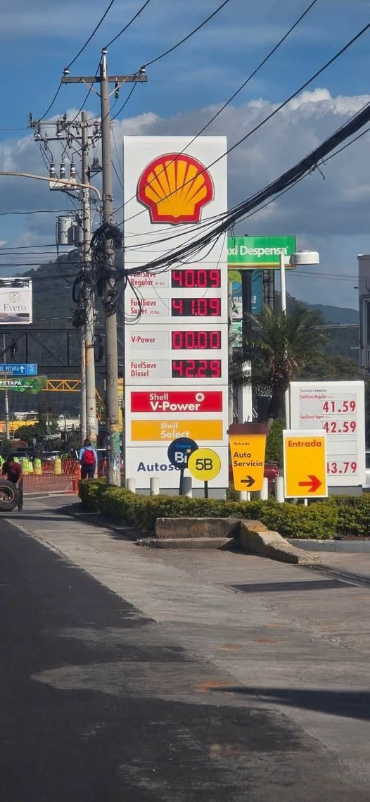 Un cartel de estación de servicio Shell exhibe los precios de distintos tipos de combustibles en una calle con postes y cables eléctricos. (Sololá Informativo Digital)