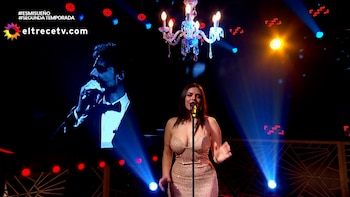 Una mujer con vestido brillante canta ante un micrófono en un escenario con luces, una lámpara de araña y una pantalla