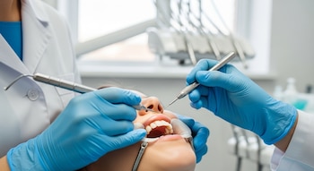Primer plano de un dentista con guantes azules examinando los dientes de un paciente con un abrebocas y herramientas dentales en una clínica.