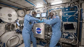 Dos astronautas con trajes espaciales ligeros, cascos y guantes manipulan un dispositivo experimental metálico con el logo de la NASA, rodeado de cables, pantallas y módulos.