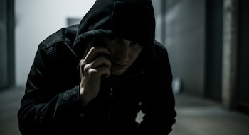 Hombre encapuchado con chaqueta oscura se lleva un teléfono móvil a la oreja. Su rostro está en sombras, visible parcialmente, en un entorno lúgubre.