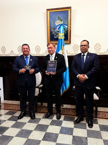 Tres hombres en trajes oscuros de pie, el de la izquierda con un documento pequeño, el del centro con un informe. Detrás, una bandera y un cuadro oficial