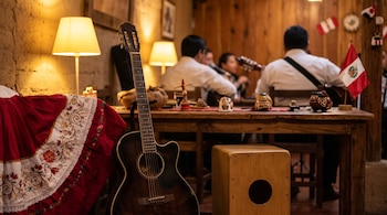 Una guitarra, un cajón de madera y una falda bordada roja junto a una mesa con figuras artesanales, una bandera peruana y músicos desenfocados.