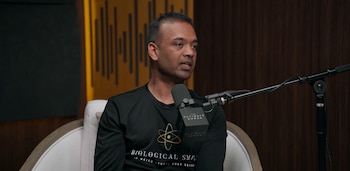 Un hombre con camiseta negra habla frente a un micrófono montado sobre un soporte en un estudio con paneles acústicos