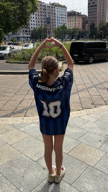 Niña de espaldas con camiseta azul y negra de Argentina con el número 10, forma un corazón con las manos, en una calle de ciudad con edificios y automóviles