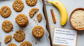 Vista superior de galletas de avena caseras en una bandeja, con un plátano, canela en rama y un cuenco de avena cruda, y una tarjeta con la receta.