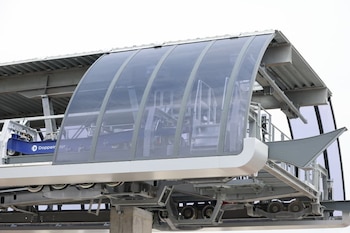 Primer plano de una sección superior de una estación de teleférico moderna con paneles curvos transparentes, estructura de metal gris y mecanismo interno visible