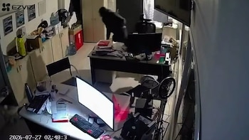 Interior de oficina con dos escritorios, monitores, sillas y gabinetes. Una persona de oscuro se mueve sobre un escritorio. Documentos, ventilador, botellas, fecha