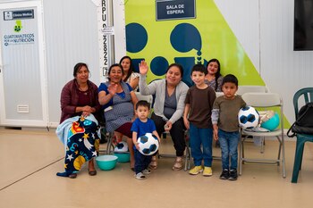 Un grupo de cinco mujeres y cuatro niños posa en una sala de espera con letreros de "Clínica Médica" y "Guatemaltecos por la Nutrición"