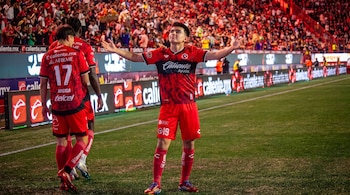 Jugador con uniforme de fútbol rojo, camiseta con escudo y pantalón corto con el número 19, con brazos extendidos. Otros jugadores, campo de juego, público y vallas publicitarias.