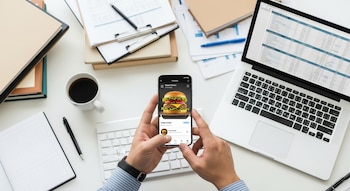 Manos de un hombre sosteniendo un celular que muestra una hamburguesa para ordenar, sobre un escritorio con documentos, laptop y taza de café.