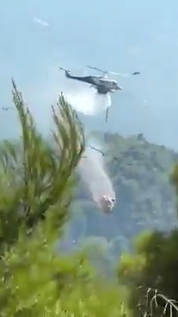 Un helicóptero vuela sobre un área boscosa y descarga agua; un segundo helicóptero cae de forma vertical a la derecha, con humo y vegetación
