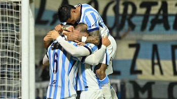 Tres jugadores de fútbol con camisetas a rayas celestes y blancas se abrazan celebrando un gol frente a una portería y una bandera