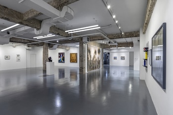 Interior de galería de arte moderna con paredes blancas y suelo gris reflectante. Muestra varias pinturas, una escultura en pedestal y techo con vigas expuestas