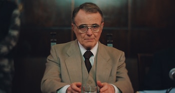 Un hombre mayor con traje beige, camisa, corbata y gafas, sentado en una mesa con un vaso de cristal. Fondo oscuro con paneles de madera