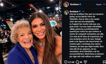 Primer plano de dos mujeres sonriendo a la cámara en un evento formal. Una mujer mayor con cabello rizado rubio y chal azul, junto a una mujer más joven con cabello castaño largo