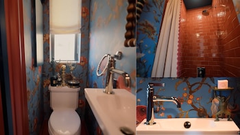 Un baño con paredes de papel tapiz floral azul y naranja, un inodoro blanco con trofeos, un lavabo moderno con grifo plateado y una ducha con azulejos rojos