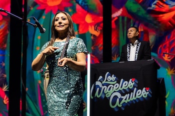 Una mujer con vestido verde de lentejuelas toca un güiro en un escenario iluminado, con un músico y el logo de Los Ángeles Azules al fondo