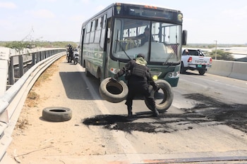 Un policía con uniforme táctico mueve neumáticos quemados de una carretera/puente, con un autobús policial y una camioneta blanca en el fondo bajo un cielo claro