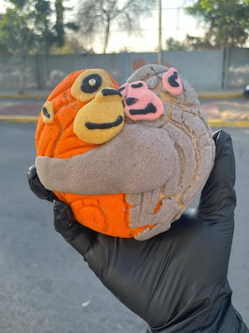 Una mano con guante negro sostiene un pan dulce tipo concha, decorado con dos caras de personajes en colores naranja y gris, con detalles amarillos y rosados