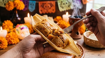 Manos sostienen un tamal abierto en hoja de maíz y un tenedor con masa, frente a velas, flores de cempasúchil y una calaverita decorada.
