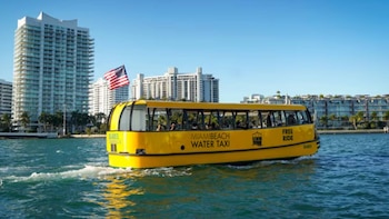 Un taxi acuático amarillo con la inscripción "Miami Beach Water Taxi" y "Free Ride" surca el agua. Al fondo, edificios altos y la bandera de EE. UU