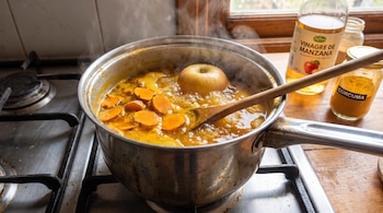 Olla de metal hirviendo con líquido amarillo, rodajas de cúrcuma y una manzana sobre estufa. Vapor, cuchara de madera, botella de vinagre de manzana y frasco de cúrcuma.