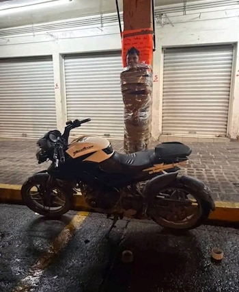 Hombre con torso y piernas envueltos en plástico transparente, atado a un poste de cemento, motocicleta blanca y negra, persianas metálicas, suelo mojado
