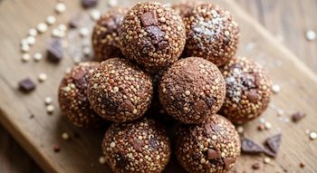 Primer plano de una pila de bocados de quinoa y chocolate caseros sobre una tabla de madera, con granos de quinoa tostados y trozos de chocolate visibles en su superficie.