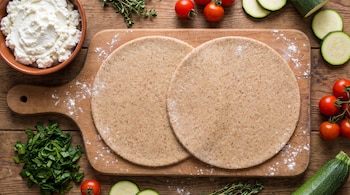 Dos discos de masa integral sobre una tabla de cortar rodeados por un recipiente con ricota, espinacas picadas, tomates cherry y calabacín.
