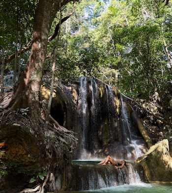 Una mujer en bikini descansa sobre una superficie de piedra junto a la caída de agua de una cascada rodeada de árboles