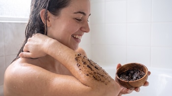 Mujer con cabello mojado se aplica exfoliante en el brazo, sostiene un cuenco de madera lleno de producto. Baño blanco de azulejos al fondo.