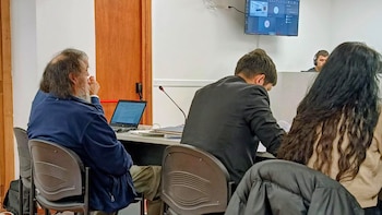 Pedro Quilodrán, un hombre de cabello y barba largos, sentado en una silla en una sala, con un portátil abierto sobre la mesa. Se ven otras personas y un monitor