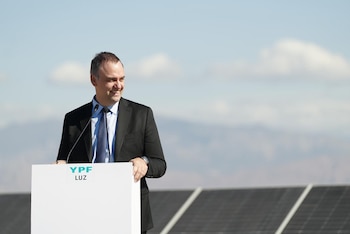 Manuel Adorni, un hombre de traje y corbata, sonríe detrás de un podio blanco con el logo 'YPF LUZ', con paneles solares y un cielo azul con nubes de fondo