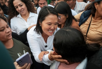 Keiko Fujimori, vestida de blanco, sonríe ampliamente mientras se inclina para interactuar con una niña de cabello oscuro en una multitud. Se ven otras personas borrosas