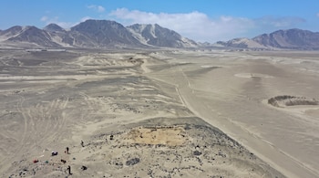 Vista aérea de una excavación arqueológica en una colina de arena con varias personas trabajando y montañas en el fondo.