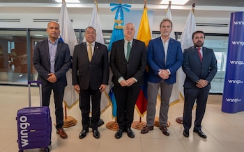Cinco hombres de traje, varias banderas, cartel azul con logo de Wingo, maleta morada con logo de Wingo y suelo claro en un interior
