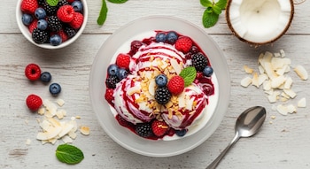 Helado sin azúcar de coco y frutos rojos en un bol blanco. Incluye frambuesas, arándanos, moras y copos de coco. En mesa de madera clara con ingredientes frescos alrededor.